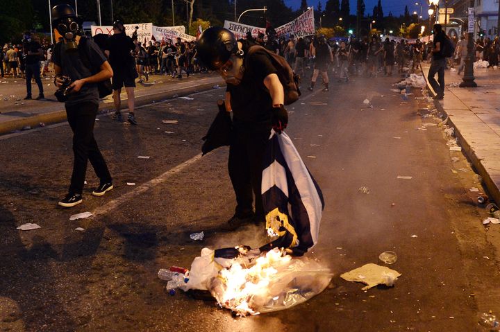 A protester burns a Greek flag in central Athens, during an anti-austerity protest on July 15, 2015. Anti-austerity protesters hurled petrol bombs at police in front of Greece's parliament on July 15, as lawmakers began debating deeply unpopular reforms needed to unlock a new eurozone bailout. Riot police responded with tear gas against dozens of hooded protesters who set ablaze parts of Syntagma square in central Athens. AFP PHOTO/ LOUISA GOULIAMAKI (Photo credit should read LOUISA GOULIAMAKI/AFP/Getty Images)