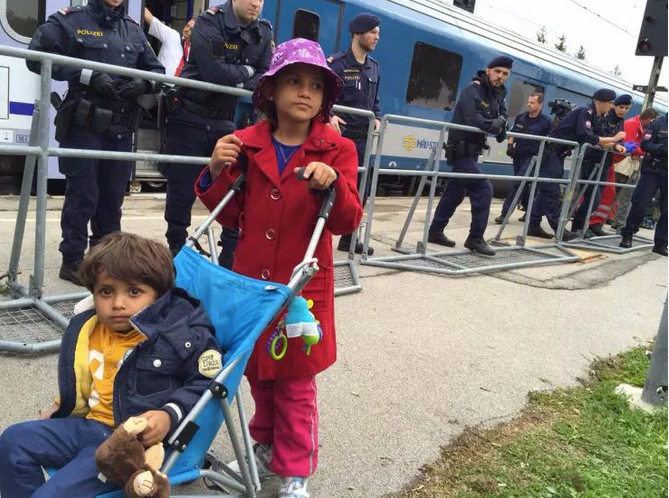 A young Syrian girl pushes her younger brother in a stroller past Austrian police guarding the train. 