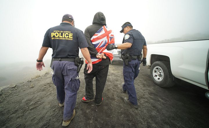 Hawaii Department of Land and Natural Resources escort a demonstrator draped in the Hawaiian flag.
