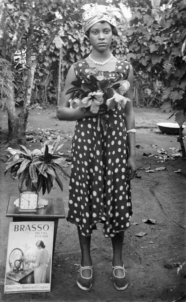 Woman standing with flowers; clock on table;"Brasso metal polish" [Dame Merry Oritsetimeyin Ehanire nee Cardigan (Osagie Ehanire's mother)]