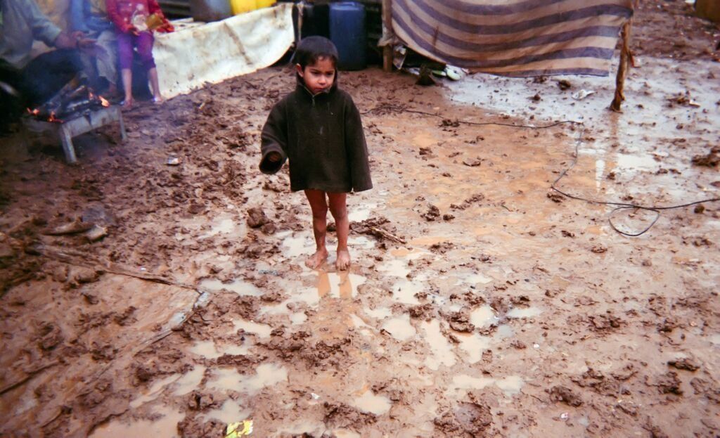 Shot by Moustafa, age 12, from Hama Governorate, Syria. A young boy who is wearing neither pants nor shoes stands on ground that is muddy from rain in the Bekaa Valley, Lebanon, in 2014.