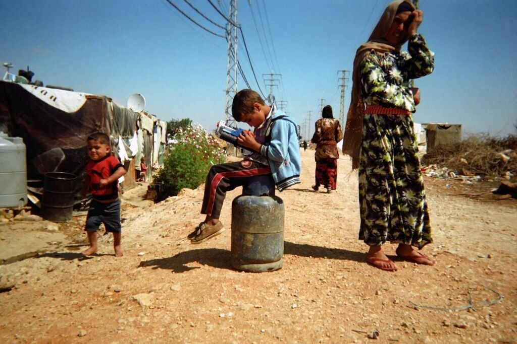 Shot by Mohamed, age 11, from Idlib Governorate, Syria. Sitting atop a metal petrol canister, a boy plays with a toy as a barefoot child runs past in the Bekaa Valley, Lebanon, in 2014.