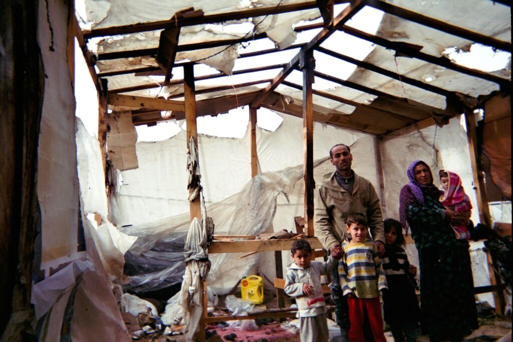 Shot by Jasem, age 7(hometown unknown).  A woman and man stand with four of their children in their makeshift shelter in the Bekaa Valley, Lebanon, in 2014. The shelter became damaged after a shoe in the stove caused a fire to break out. This is the photographer's family.