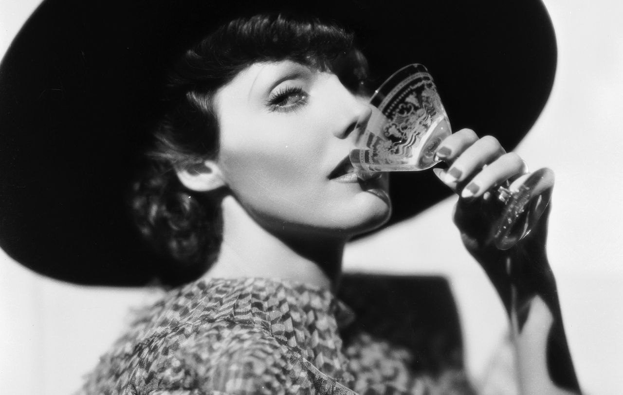 American actress Adrienne Ames circa 1930, dressed in a chiffon tiered dress, drinks from a cocktail glass.