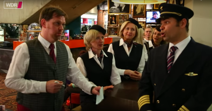 Singers dressed like a pilot and a waiter are seen breaking into song among several flight attendants who appear stunned.