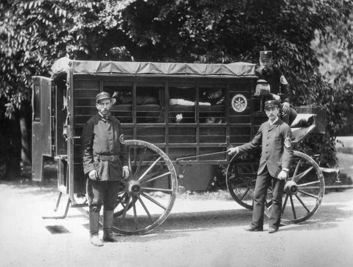 A Wiener Ambulance with patients in 'layers' in a horse drawn wooden carriage. The sides are partly open, but have curtains. The ambulance men are members of the Viennese Voluntary Rescue Society founded in 1881.