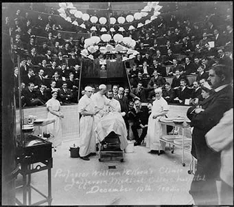 Surgeons gather around the operating table in the clinic amphitheater at the Jefferson Medical College Hospital, with spectators and medical students seated in the background, 1902.