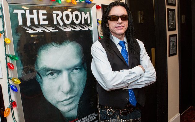 Tommy Wiseau poses in front of a poster for "The Room."