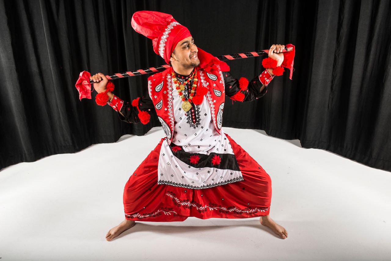 CU Bhangra dancer Ishaan Kolluri squats low, with a large wooden stick known as a "koonda" balanced on his shoulders.