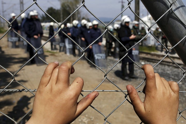 A child places his hands on a fence as Greek police officers stand guard after minor clashes occurred at a makeshift camp for migrants and refugees at the Greek-Macedonian border near the village of Idomeni.