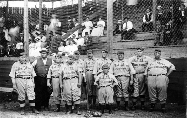 Players on The Fat Man's Baseball Association, wear jerseys written with "Fat Mens Amusement" across it and pose in their home ballpark (unknown location) in 1910. 