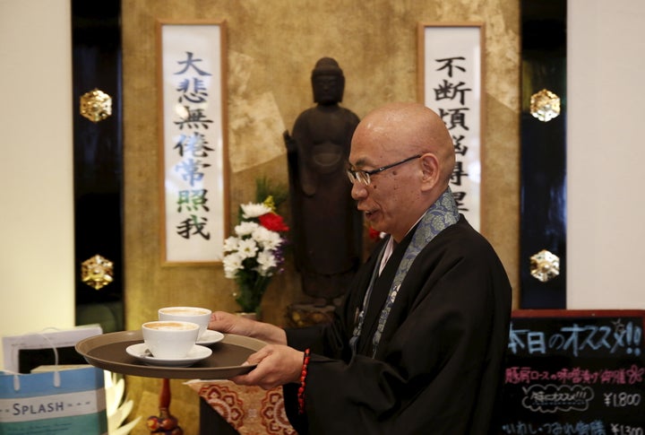 Shokyo Miura, a Buddhist monk and one of the on-site priests, carries cups of coffee past a statue of Buddha at Tera Cafe in