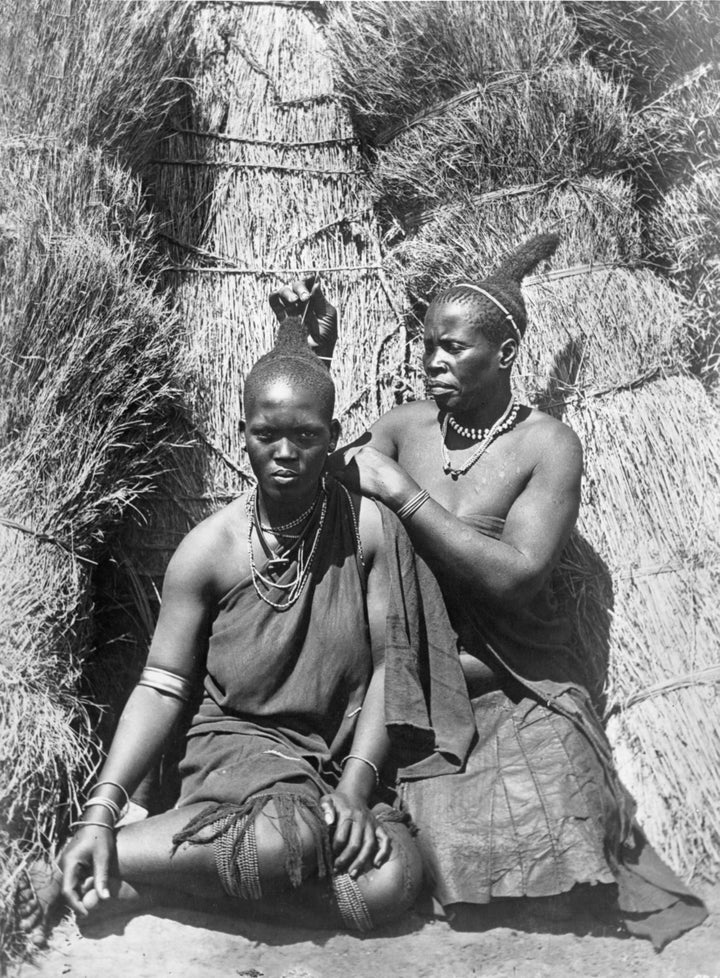 An adult Zulu woman grooms and adorns the hair of a younger woman in South Africa.