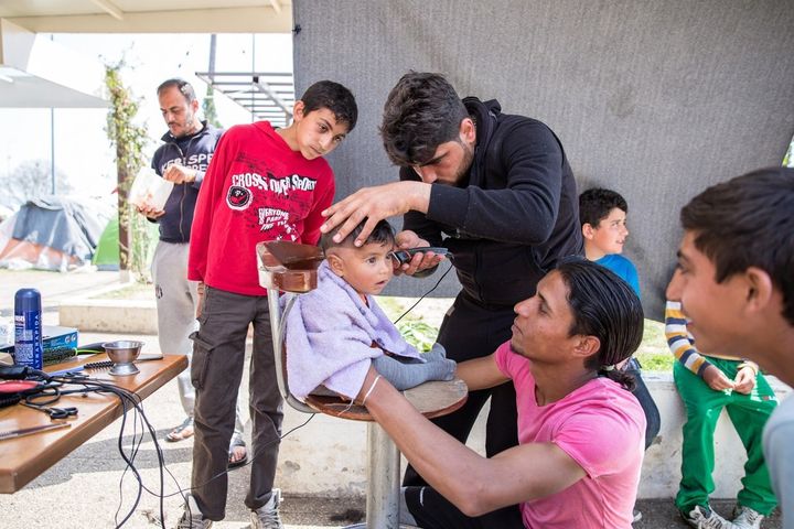 Nine-month-old Ahmad, from Syria, gets a haircut while his 23-year-old father, Farhud, holds his attention. Ali, the barber, from the Syrian city of Aleppo, was a women's hair stylist, but now mostly offers his services to young men in the camp, charging 5 euros for a haircut and shave.