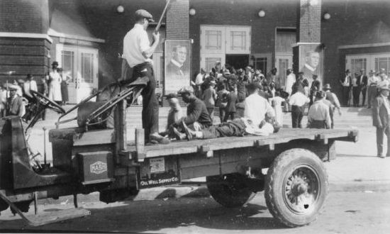 A victim of the Tulsa race riot lays on the back of a flat bed truck outside Convention Hall as a white man with a shotgun stands on the truck.