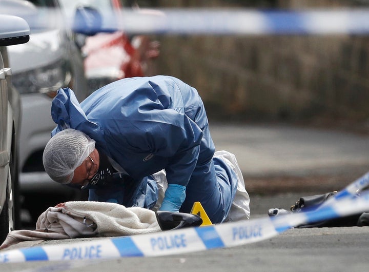 A forensics police officer works next women's shoes and a handbag on the ground behind a police cordon