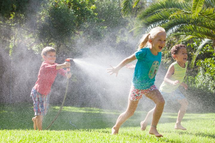 Three children in garden chasing each other with water sprinkler RUSS ROHDE via Getty Images