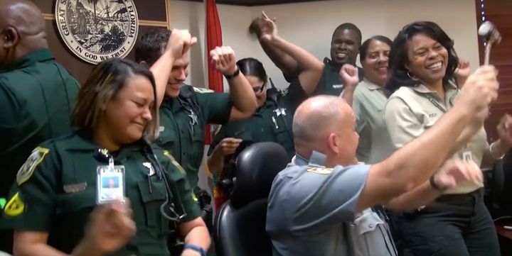 A uniformed man is waves around a judge's gavel as his companions dance wildly around him.