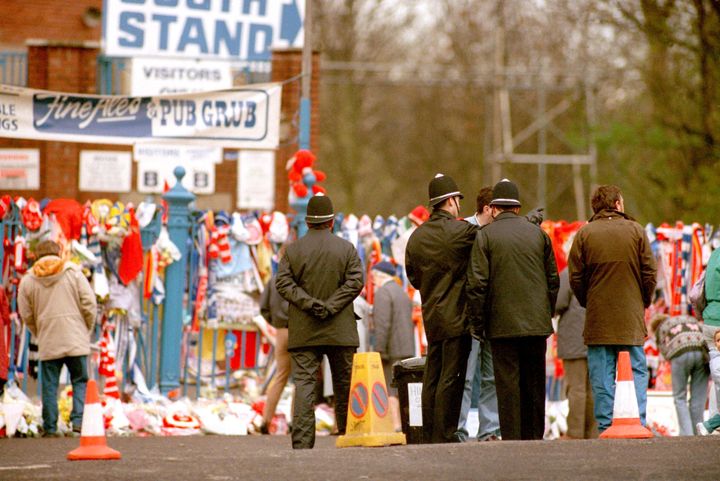 Policemen stand outside the Leppings Lane end of the Hillsborough ground, as the flowers pile up in memory of the fans that died
