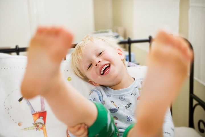 Selective focus view of toddler boy laying in bed smiling, holding his feet up to the viewer. He is in pajamas with barefeet and clown sheets. AE Pictures Inc. via Getty Images
