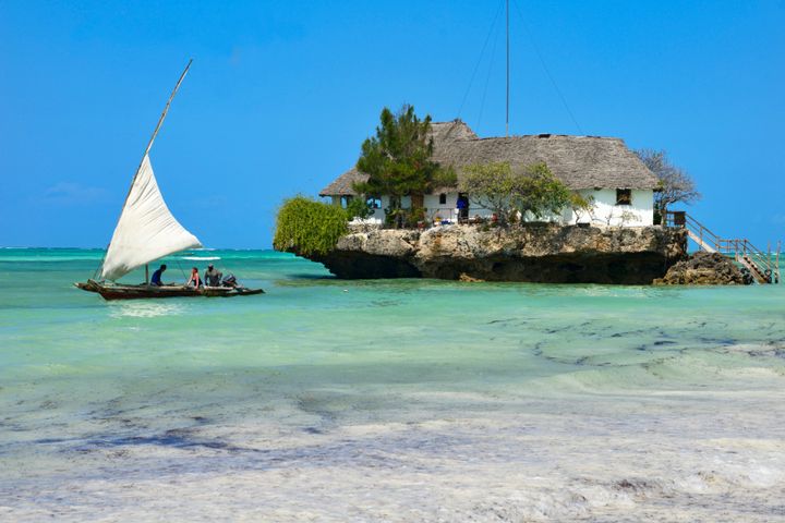 Tourist take a traditional Dhow boat to The Rock Restaurant just off Zanzibar's idyllic Bwejuu Beach.