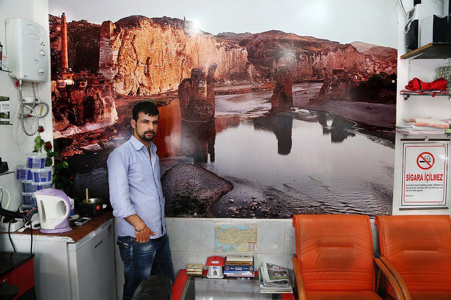 Adar, a hairdresser, poses in front of a poster of Hasankeyf that hangs on the wall of his salon.