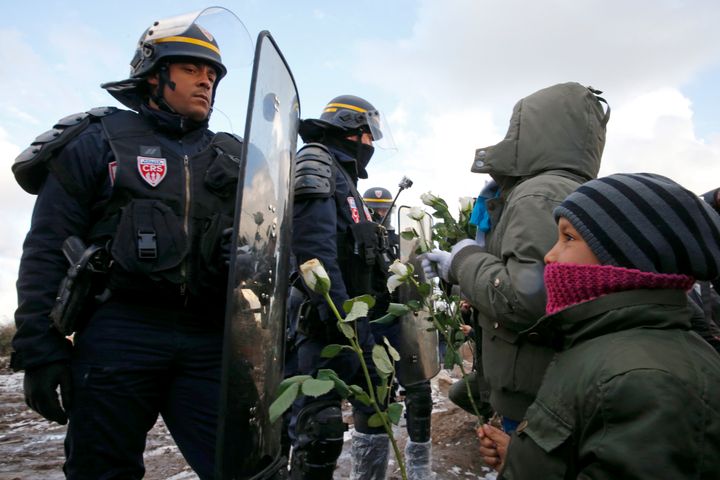Young migrants holding white flowers face French riot police officers who secure the area near makeshift shelters in Calais, France, March 7, 2016.