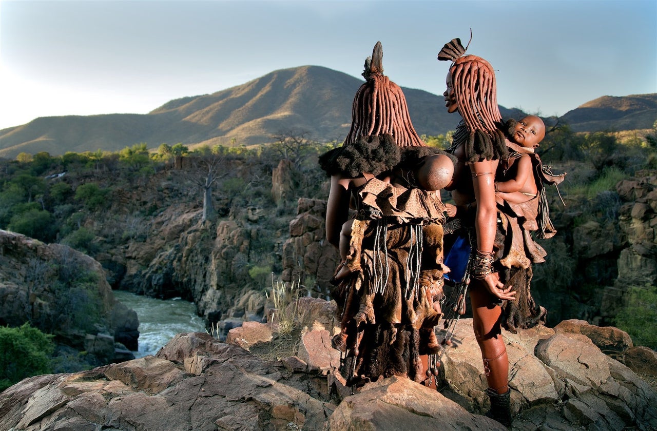 Young mothers carry their babies in traditional carriers looking over the Kunene river. Himba tribe, Namibia.