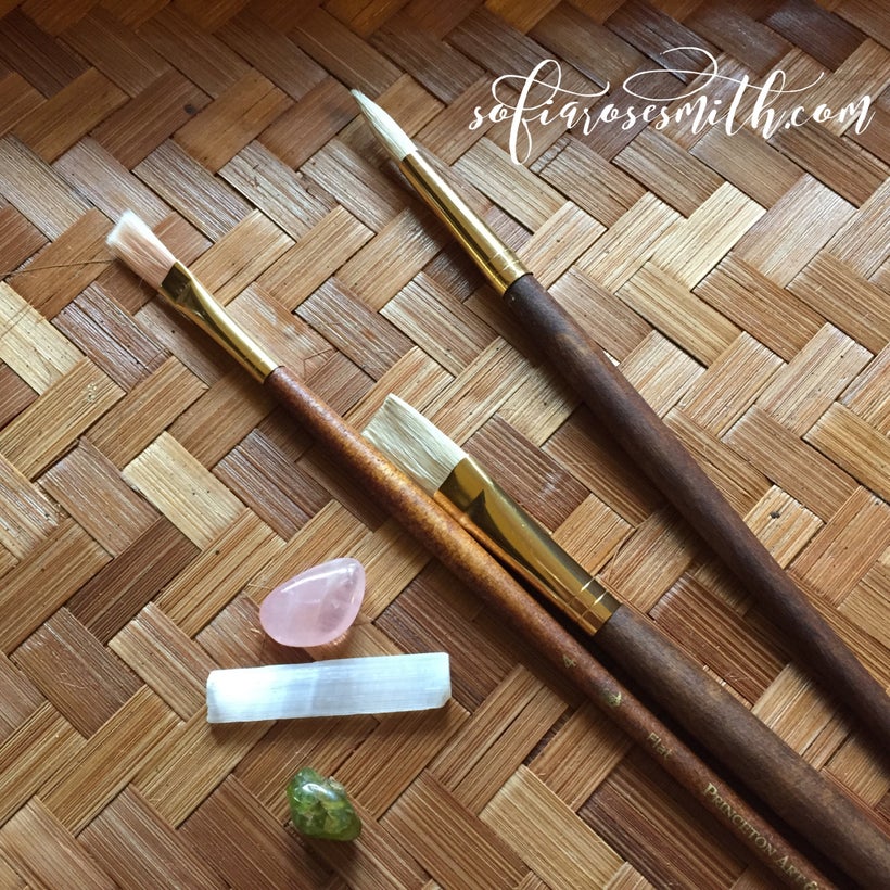 three wooden paint brushes lay atop a bamboo tray. a few stones sit beside them: rose quartz, selenite, and peridot. the text