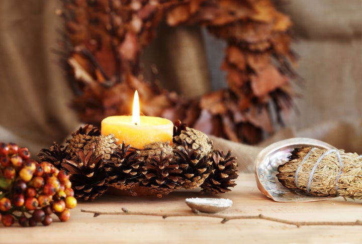 A pagan altar is decorated for Samhain. Altar items include a sage smudge stick resting in an abalone shell, a hazel wand, a metal bowl of salt, a candle and various natural items.