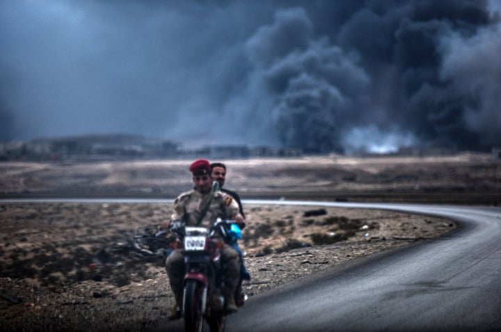 An Iraqi soldier and a civilian ride a motorbike as smoke rises behind them, on the road between Qayyarah and Mosul on Octobe