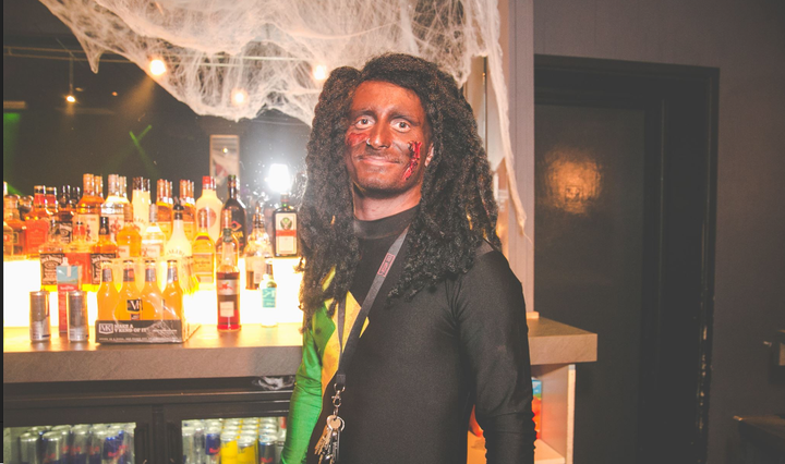 The photo shows a member of staff in blackface an afro wig and the colours of the Jamaican flag