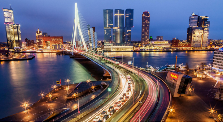 <p><em>Evening view of Rotterdam skyline and waterfront with the suspension cables of the city's emblematic Erasmus Bridge illuminated against the sky.</em> </p>