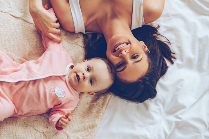 Top view of cheerful beautiful young woman looking at camera with smile while lying in bed with her baby girl g-stockstudio via Getty Images