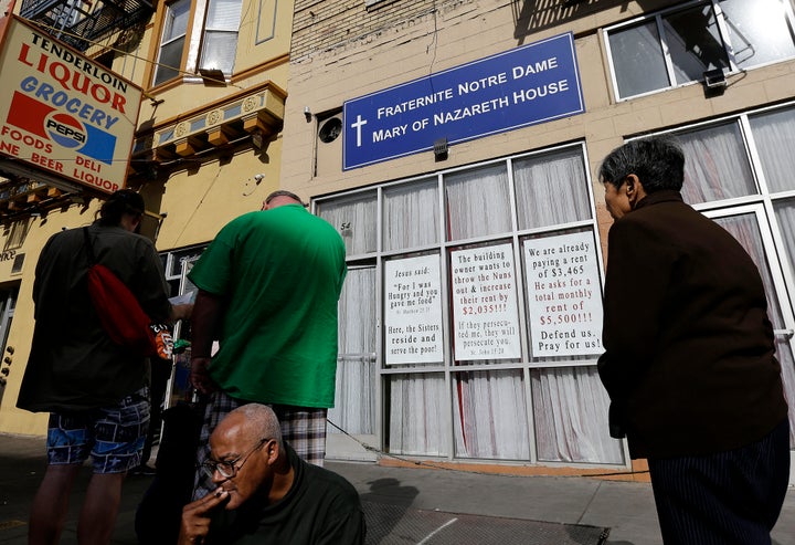 Signs hang on the front of the Fraternite Notre Dame Mary of Nazareth Soup Kitchen behind a line of people waiting for food t