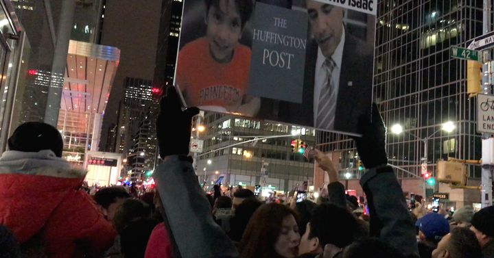 Two fans of Isaac give each other a New Year Kiss under the poster of Isaac's article at Times Square at 12:01 am, 2017