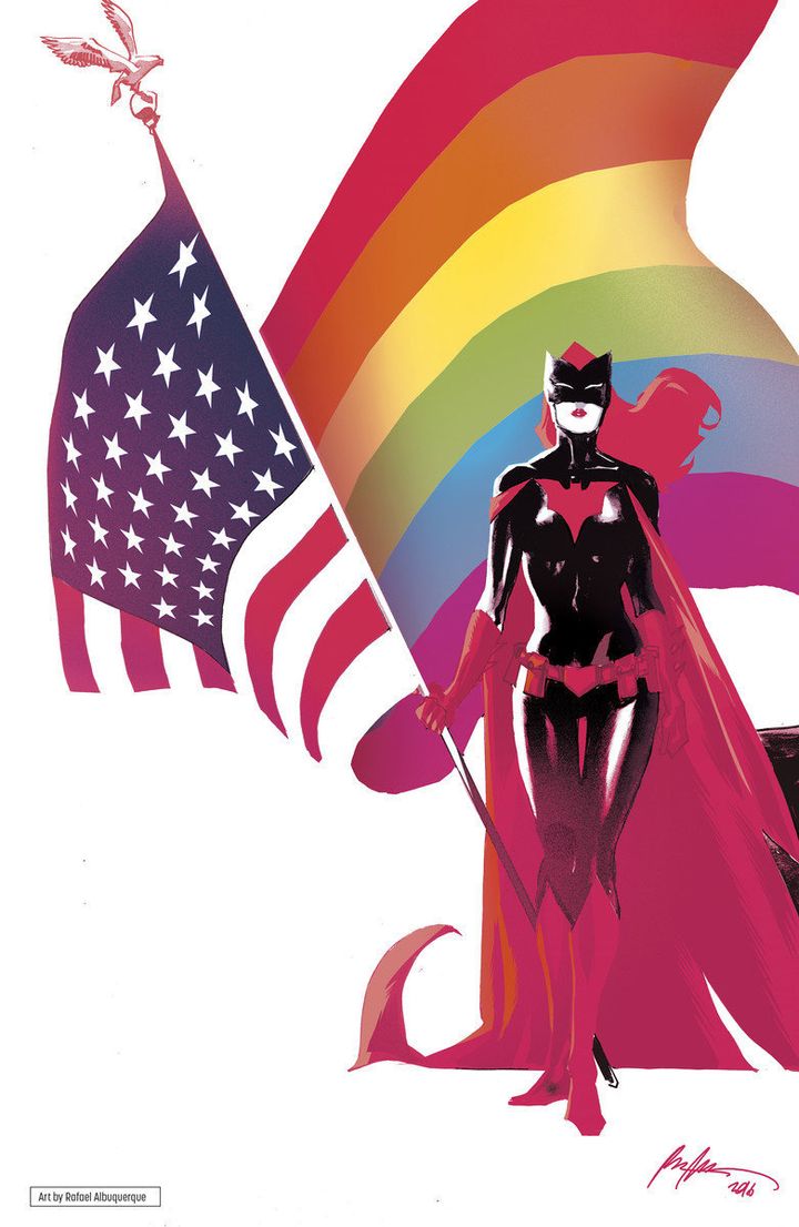 Batwoman holds an American flag with an LGBTQ flag waving behind her.