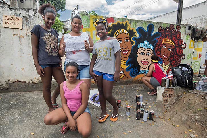 AMO Crew from Brazil. Carla Felizardo – NEGRA, Lu Brasil, Mariana Maia – Ato and Taina Xavier- Baker painted the portraits of these girls on the wall of their home. Their names are, Ana Luiza, Laryssa and Marcelly. The girls’ grandmother had wished for portraits of her granddaughters painted during last year’s event but sadly she passed away. This year the girls’ mother, Ms. Gomes shown here holding the sketch for the mural arranged to fulfill the grandmother’s dream with the help of the AMO Crew. Meeting Of Favela 2016. Favela Operaria. Duque de Caxias. Rio De Janeiro, Brazil. 