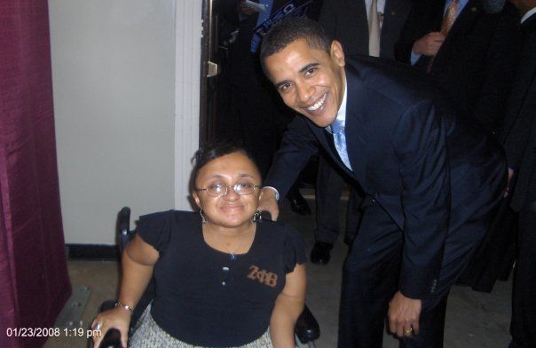 Image of me with then Sen. Obama in 2008. In the photo is a young light-skinned Black woman in a wheelchair with a tall light-skinned Black man leaning next to her. Both people are smiling for the camera. 