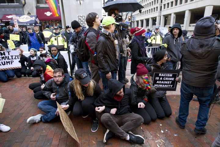 Demonstrators sit on sidewalk attempting to block people entering a security checkpoint.