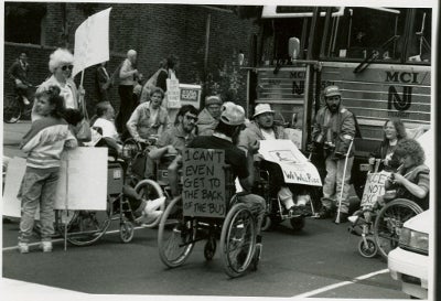 People with a range of disabilities publicly protesting in front of a bus in the 1970s, before the Americans with Disabilities Act was passed. One man in a wheelchair has a sign that reads, “I can’t even get to the back of the bus.”