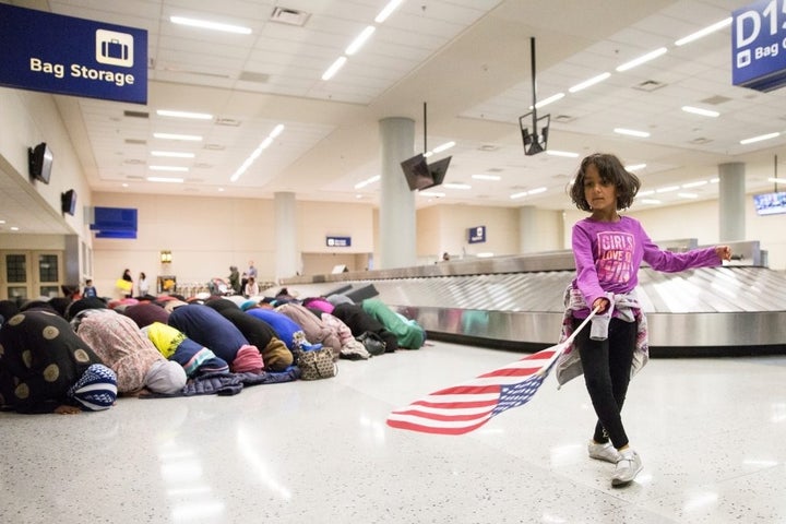 A young girl dances with an American flag while women pray behind her during a protest of the temporary travel ban .