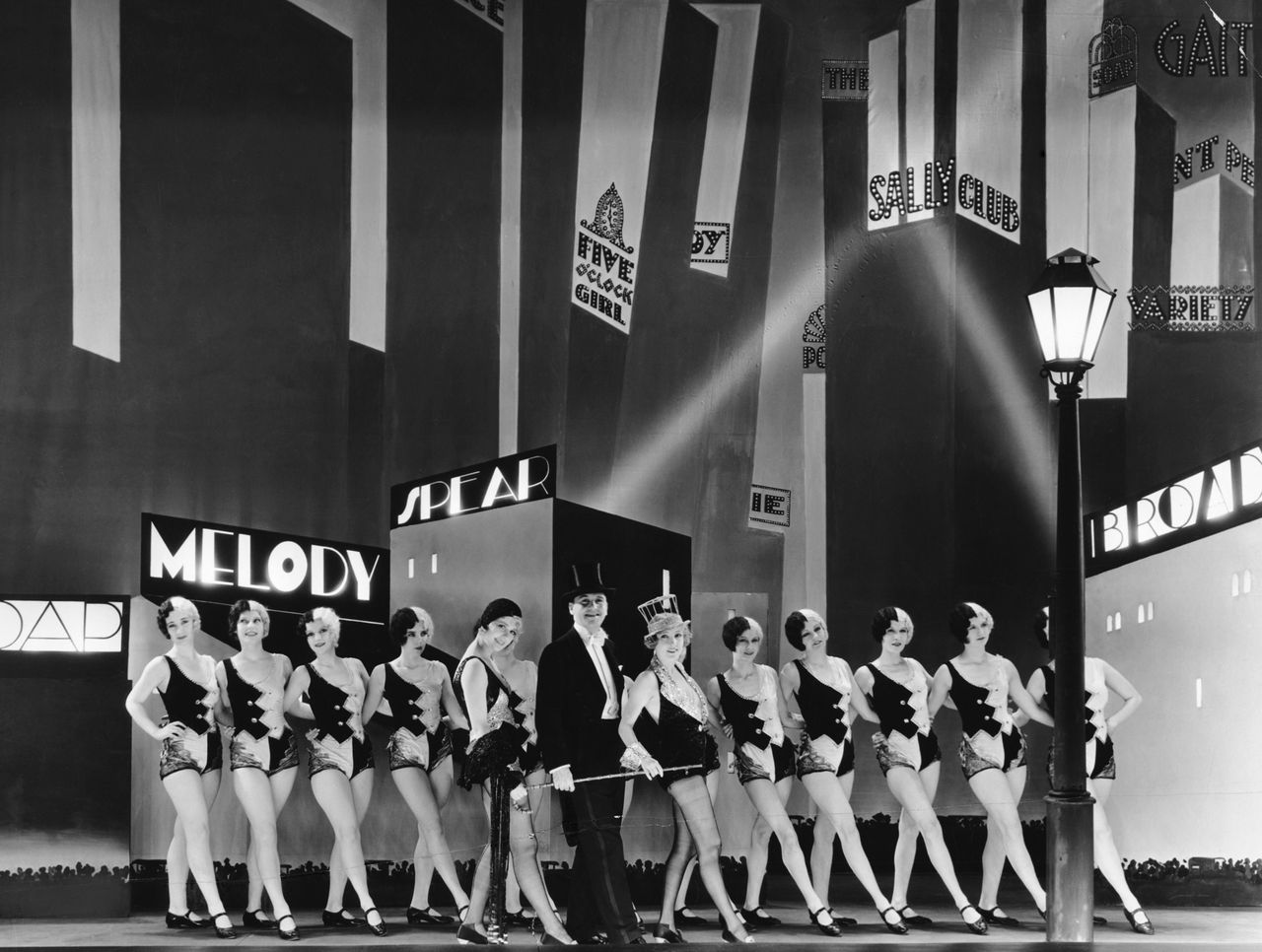 Charles King and Anita Page, stars of the film, stand with dancers in front of a Manhattan backdrop.