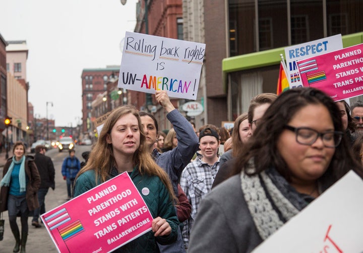 A group of people walk down a city street holding signs, the most prominent of which reads “Rolling back rights is Un-American!”