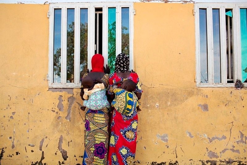 Women watch as people carry food supplies out of a storage building at the Fufore camp for internally displaced persons outsi