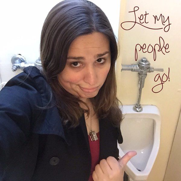 Hannah Simpson selfies before a urinal, with wall writing, “Let My People Go!” in protest of transphobic bathroom laws.