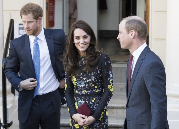 Prince Harry and the Duchess & Duke of Cambridge in London earlier this year. Harry is wearing a suit with a blue tie, the Duchess is wearing a black dress with grey and yellow flowers, and the Duke is wearing a suit with a maroon tie.