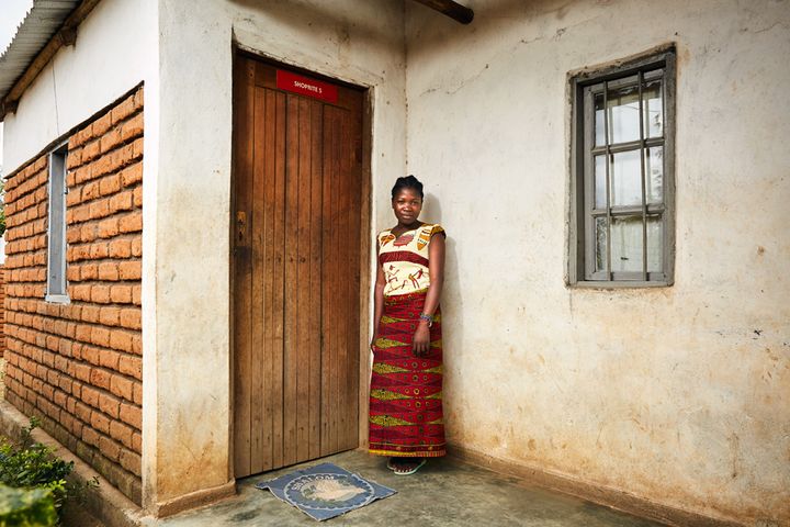 Happiness (17) stands next to the front door of her gowelo, Thyolo District, Malawi, 2016. She has lived here since 2015. Like all the others, her gowelo has a living room, a bedroom and a storage room. Her father died when she was very young, and she was sent to an orphanage by her mother, where she remained until she was brought back to her village. Happiness now lives opposite her mother, in the community where she was born. 