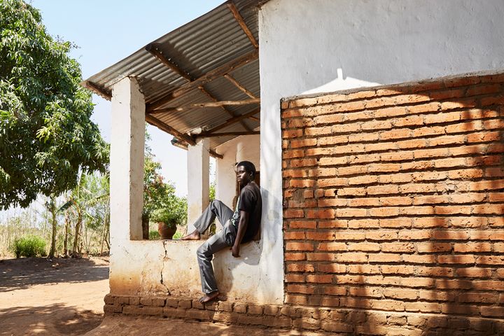 James (19) sits on the porch of his gowelo, Thyolo District, Malawi, 2016. James was very young when his father died. Although his mother is still alive, she is unable to support him as women in Malawi have a far harder time finding employment than men. James is proud of his gowelo, which he decorated himself. When he is not studying to become a plumber, he likes to listen to music or the news on a portable radio which he powers with a car battery. His dream is to one day open his own business.