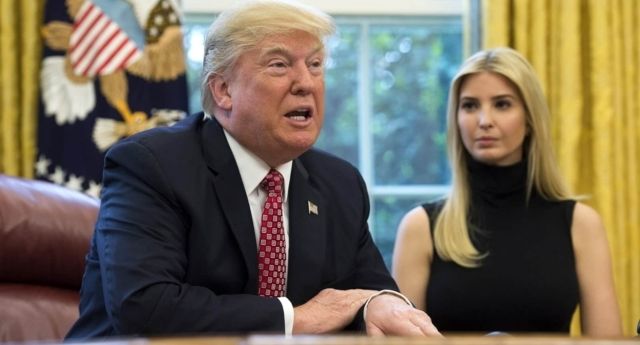 President Trump is in the foreground and his daughter Ivanka behind him to his right. He is wearing a suit with a red tie and she is wearing a black turtleneck.